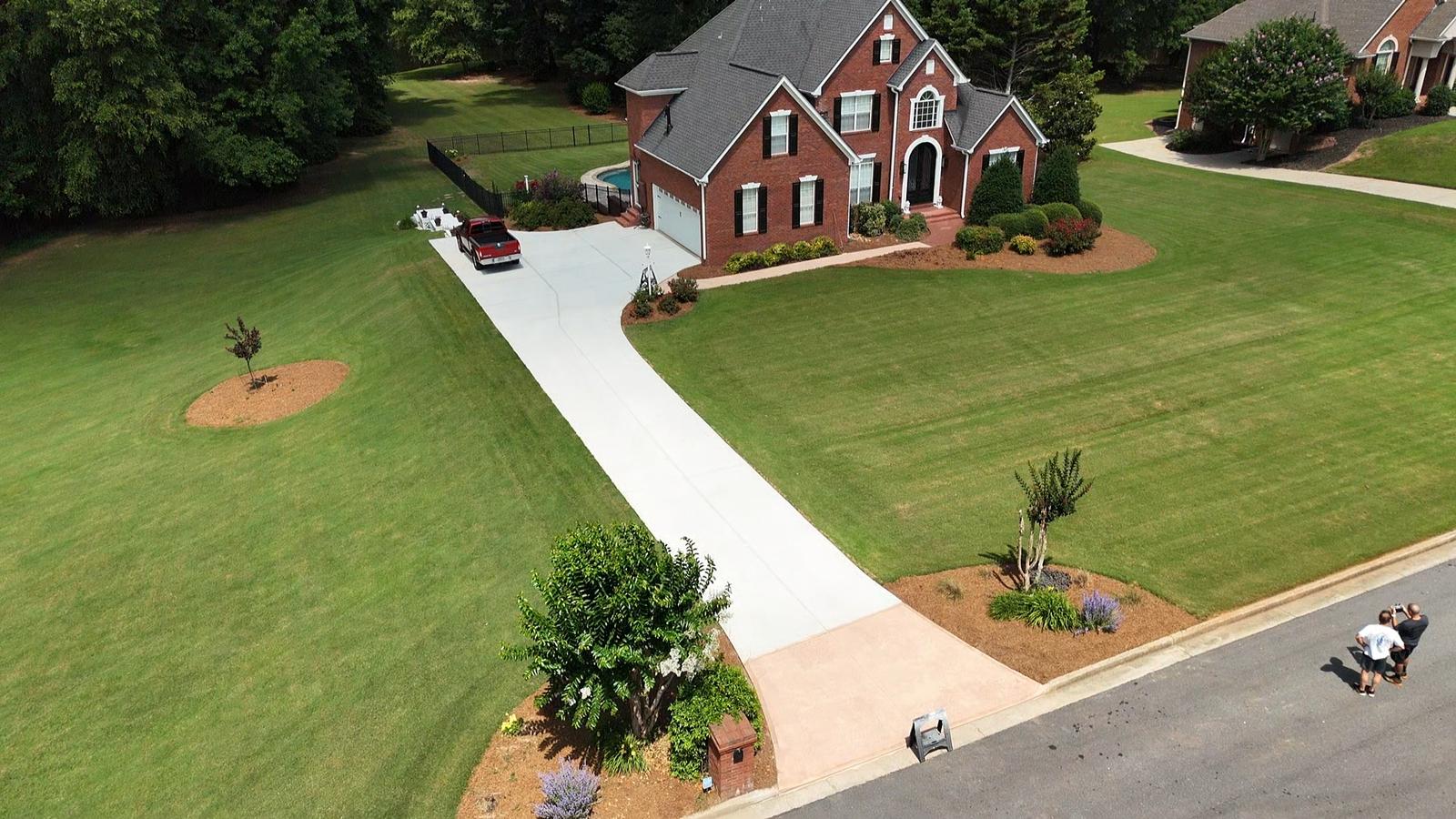 Aerial view of a suburban house showcasing a newly installed white concrete driveway by Boss Concrete. The two-story brick home features black shutters and a prominent front porch, surrounded by a well-manicured lawn. A red pickup truck is parked nearby, while circular flower beds with small trees and vibrant flowers enhance the landscape. The lawn is lush and green, with two individuals on the right side of the image engaged in conversation.