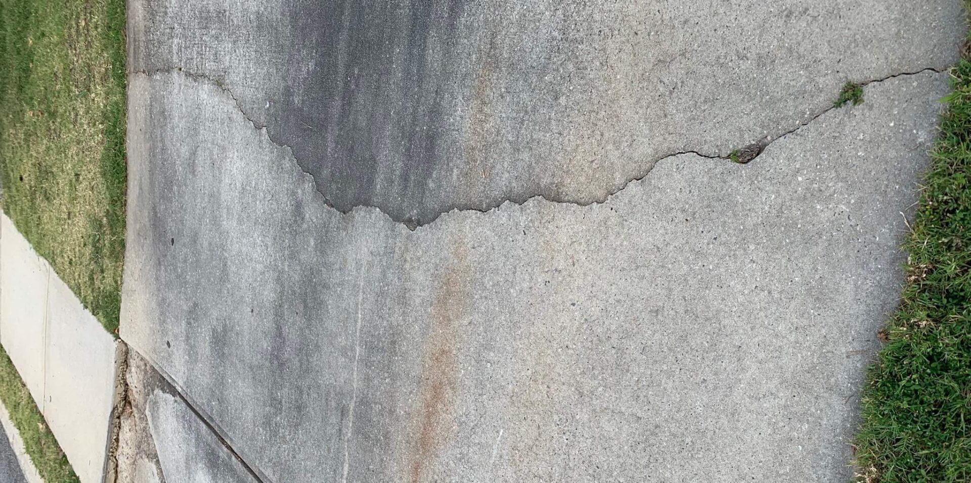 Cracked concrete driveway leading to a sidewalk with a patch of grass beside it. The driveway shows discoloration and wear, while the sidewalk connects to the curb near the road.